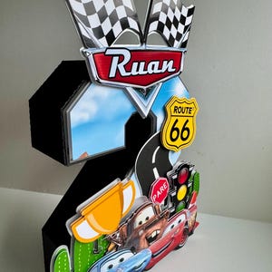 May include: A black number two birthday decoration featuring Cars characters, Route 66 signage, and checkered flags. The decoration includes the name "Ruan" and a road with a traffic light.