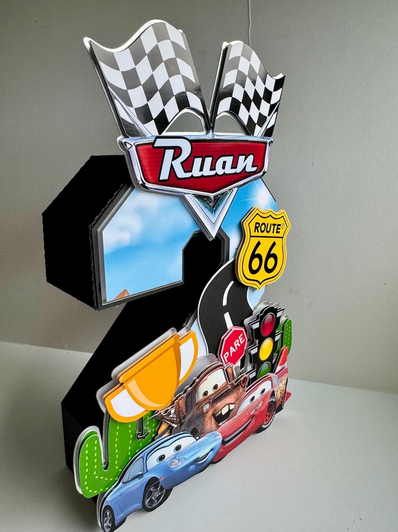 May include: A black number two birthday decoration featuring Cars characters, Route 66 signage, and checkered flags. The decoration includes the name "Ruan" and a road with a traffic light.