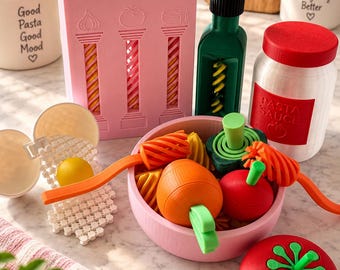 Op de afbeelding: Een roze rechthoekige pastapers met drie kolommen, een groene fles, een witte pot met het label PASTA SAUCE en een rood deksel, en een roze kom met oranje en rood speelgoedgroente. Een wit eivormig bakje met een gele dooier is aanwezig.