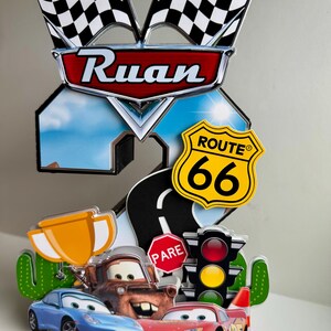 May include: A colorful cake topper featuring the name "Ruan" above a number "2" with a Route 66 sign and checkered flags. The design includes cartoon cars, a trophy, and a stop sign.