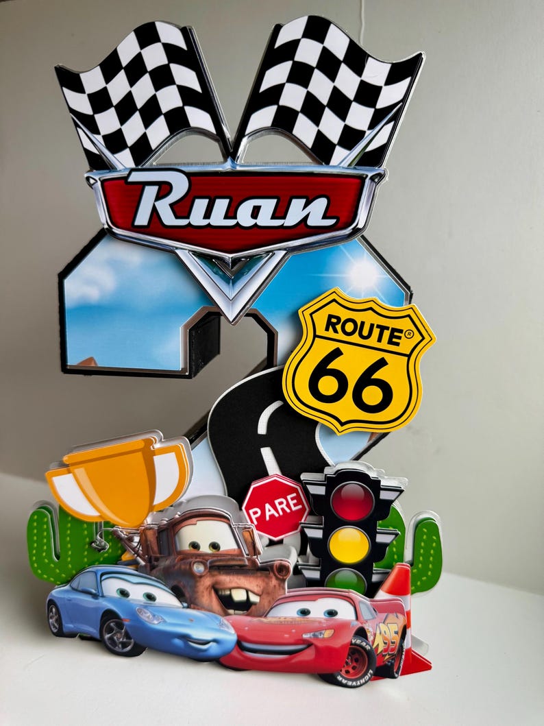 May include: A colorful cake topper featuring the name "Ruan" above a number "2" with a Route 66 sign and checkered flags. The design includes cartoon cars, a trophy, and a stop sign.