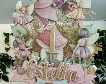 May include: A whimsical cake topper featuring a fairy theme. The design includes fairies, a mushroom, and the number "1" in gold. The name "Estella" is displayed on a pink banner, with glitter accents.