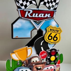 May include: A colorful cake topper featuring characters from the movie Cars, including Lightning McQueen, Mater, and Sally. The design includes a Route 66 sign, checkered flags, and the name "Ruan".