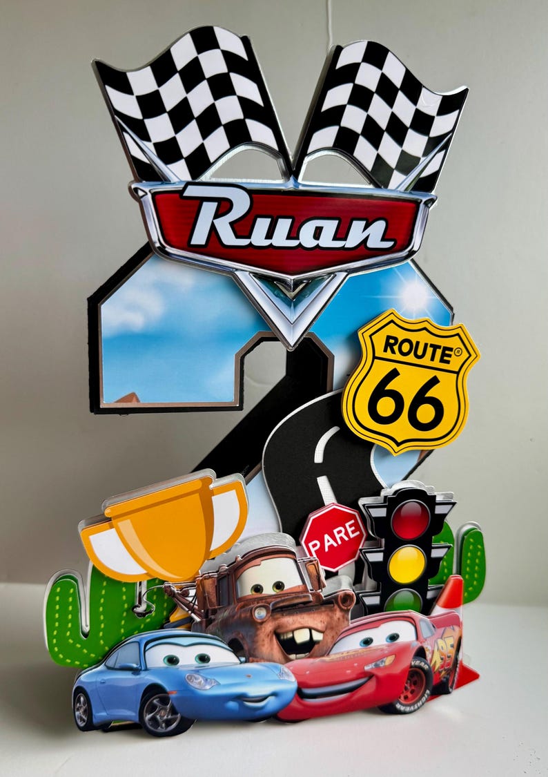 May include: A colorful cake topper featuring characters from the movie Cars, including Lightning McQueen, Mater, and Sally. The design includes a Route 66 sign, checkered flags, and the name "Ruan".
