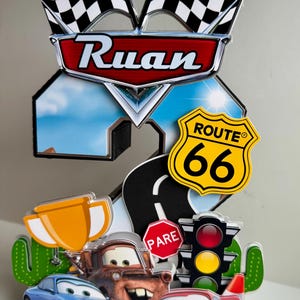 May include: A colorful birthday cake topper featuring the name "Ruan" in a red and silver emblem, with checkered flags, Route 66 sign, and cartoon cars. Includes a road, stop sign, traffic light, and a trophy.