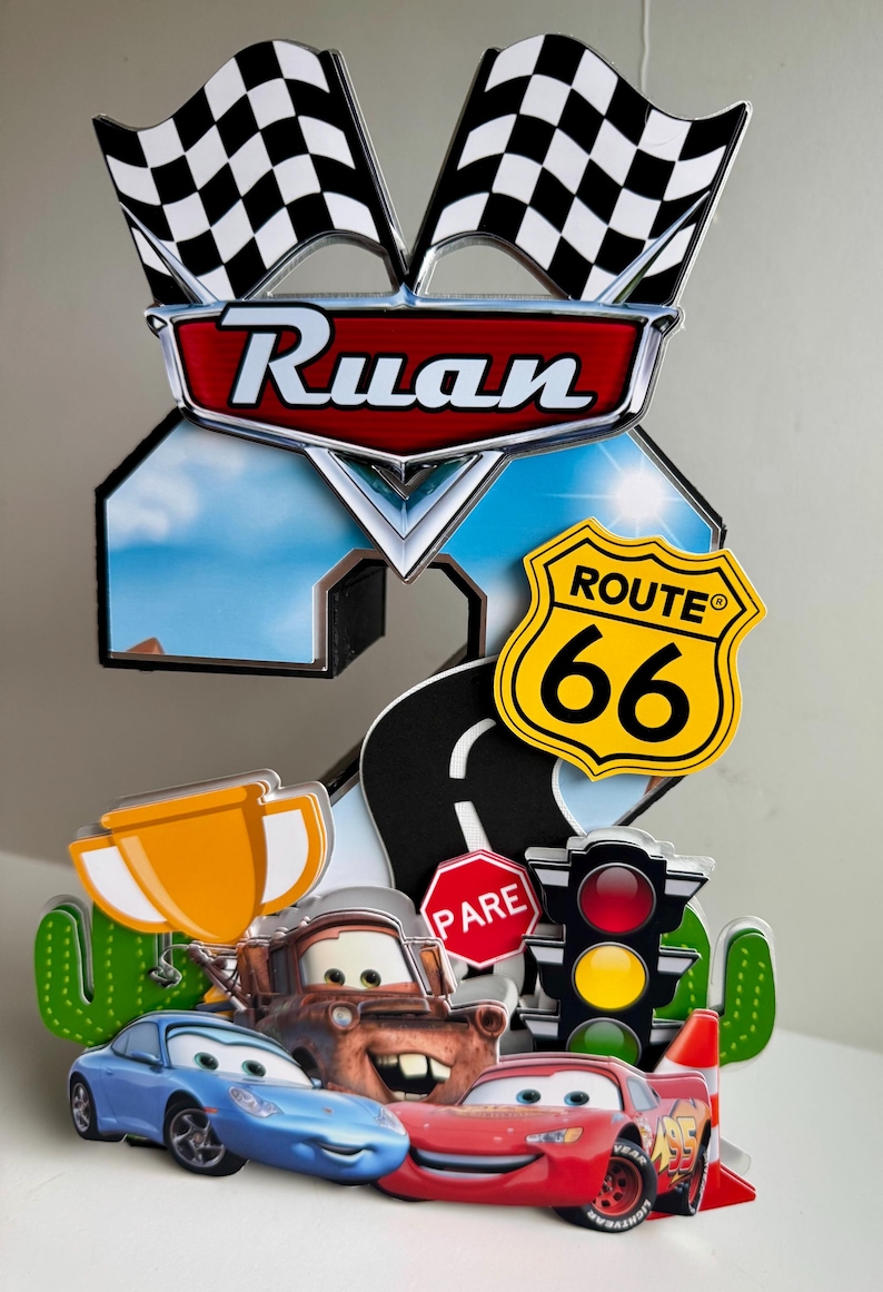 May include: A colorful birthday cake topper featuring the name "Ruan" in a red and silver emblem, with checkered flags, Route 66 sign, and cartoon cars. Includes a road, stop sign, traffic light, and a trophy.