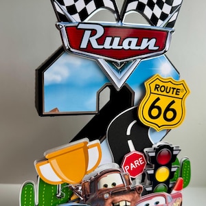 May include: A colorful cake topper featuring characters from the movie Cars, including Lightning McQueen, Mater, and Sally. The topper includes a Route 66 sign, checkered flags, and the name "Ruan".
