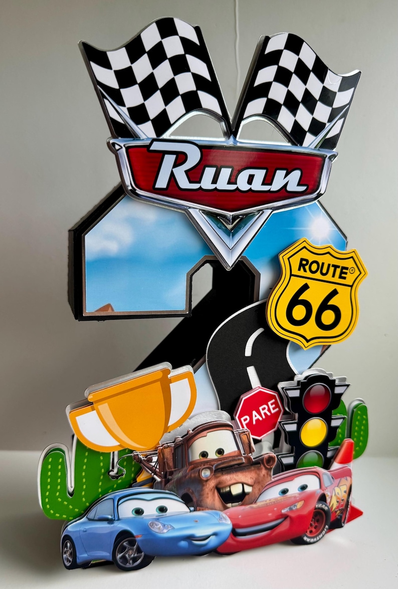 May include: A colorful cake topper featuring characters from the movie Cars, including Lightning McQueen, Mater, and Sally. The topper includes a Route 66 sign, checkered flags, and the name "Ruan".