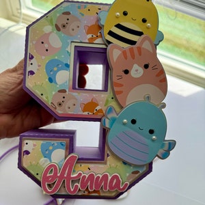 May include: Purple number nine cake topper with a pastel background and various animal cutouts. The animals include a bee, a cat, a ladybug, and a penguin. The topper also includes the name "Anna" in pink letters.