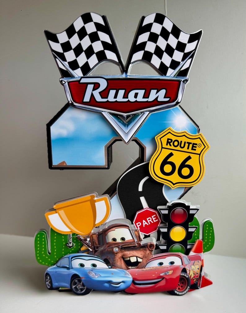 May include: A colorful birthday cake topper featuring the name "Ruan" in a red and silver emblem, with checkered flags, Route 66 sign, and cartoon cars. Includes a trophy, stop sign, and traffic light.
