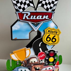 May include: A colorful cake topper featuring characters from the movie Cars, including Lightning McQueen, Mater, and Sally. The topper includes a Route 66 sign, checkered flags, and the name "Ruan".