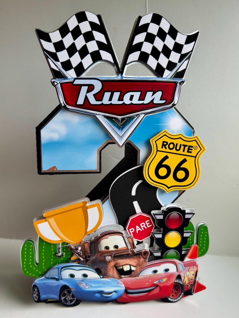 May include: A colorful cake topper featuring characters from the movie Cars, including Lightning McQueen, Mater, and Sally. The topper includes a Route 66 sign, checkered flags, and the name "Ruan".