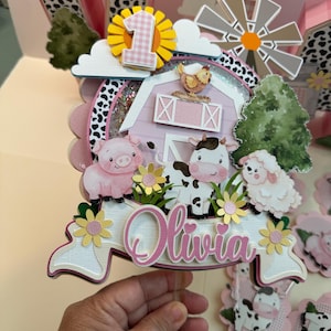May include: A pink and white farm-themed cake topper with a barn, windmill, clouds, flowers, and animals. The topper says "Olivia" and has the number "1" on it.