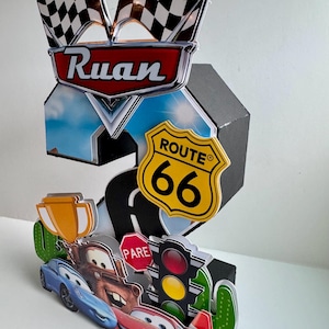 May include: A colorful, layered birthday decoration featuring a racing theme. It includes checkered flags, the name "Ruan", Route 66 signage, and cartoon car characters. The design incorporates a road, traffic lights, and a trophy.