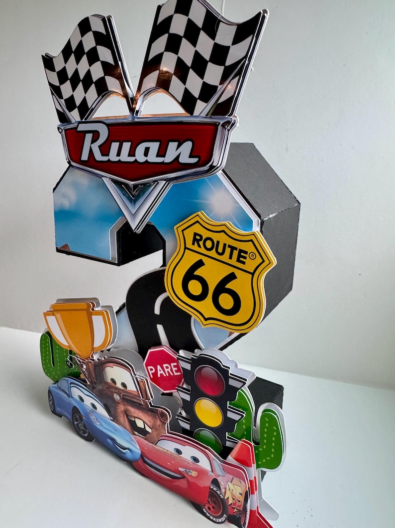May include: A colorful, layered birthday decoration featuring a racing theme. It includes checkered flags, the name "Ruan", Route 66 signage, and cartoon car characters. The design incorporates a road, traffic lights, and a trophy.