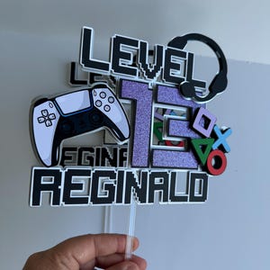 May include: A cake topper with a white and black video game controller, a purple glittery number 15, a black and white headset, and the text "Level 15 Reginald".