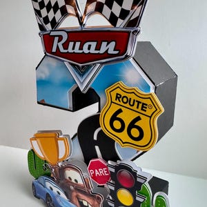 May include: A 3D papercraft featuring the name "Ruan" above a Route 66 sign and checkered flags. The design includes cartoon cars, a stop sign, and a traffic light, all set against a sky backdrop.