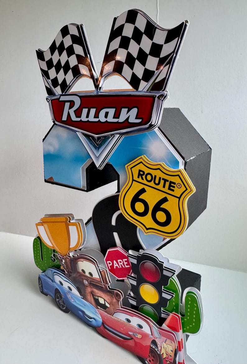 May include: A 3D papercraft featuring the name "Ruan" above a Route 66 sign and checkered flags. The design includes cartoon cars, a stop sign, and a traffic light, all set against a sky backdrop.