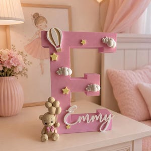May include: A large, pink, three-dimensional letter E featuring the name Emmy in white cursive script. The letter is decorated with a small beige teddy bear holding a cluster of balloons, a hot air balloon, several yellow stars, and white clouds.