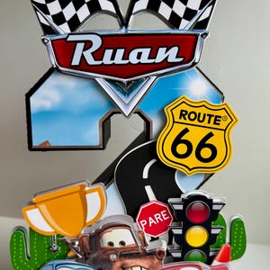 May include: A colorful cake topper featuring the name "Ruan" with racing flags, Route 66 sign, and cartoon cars. Includes a trophy, stop sign, and traffic light. The design is vibrant and playful.