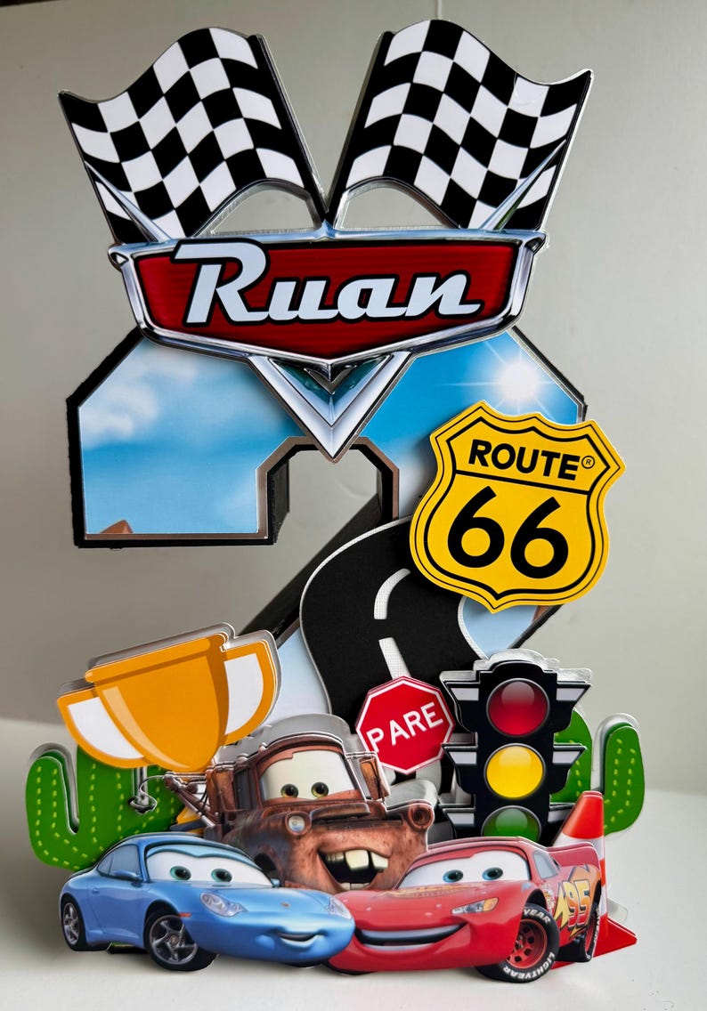 May include: A colorful cake topper featuring the name "Ruan" with racing flags, Route 66 sign, and cartoon cars. Includes a trophy, stop sign, and traffic light. The design is vibrant and playful.