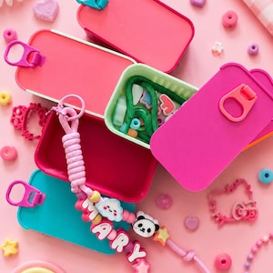 May include: A collection of rectangular plastic containers in shades of pink and teal, each featuring a pull-tab lid. A beaded keychain with the name MARY, a panda charm, and a cloud charm rests on top of the open containers filled with green cord.