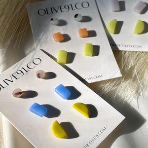 Op de afbeelding: Drie sets kleurrijke geometrische oorbel studs op witte kaartjes. De kaartjes hebben de tekst "OLIVE91.CO" en "OLIVE91CO.ETSY.COM". De oorbel studs zijn in de vorm van cirkels, vierkanten en halve cirkels. De kleuren omvatten geel, blauw, oranje en wit.