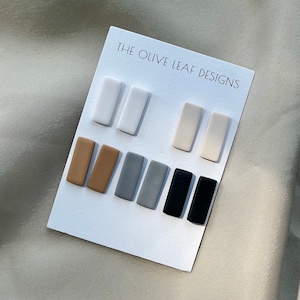 May include: A set of eight rectangular ceramic earrings in various colors, including white, beige, brown, gray, and black. The earrings are displayed on a white card with the text "THE OLIVE LEAF DESIGNS".