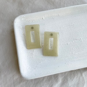 May include: A pair of light green rectangular earrings with a small opening in the center. The earrings have a speckled texture.