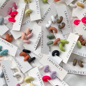 Könnte beinhalten: Sortiment an bunten, handgefertigten Polymer Clay Creolen. Die Ohrringe werden auf weißen Karten mit dem aufgedruckten Text "OLIVE91.CO" präsentiert.