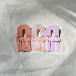 以下が含まれることがあります： 手作りのポリマークレイ製のイヤリングが3ペア。イヤリングはアーチ型で、長方形のものがぶら下がっています。色はピンク、白、薄いパープルです。