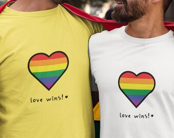 May include: Two people wearing t-shirts with a rainbow heart and the text "love wins!" printed on them. They are standing in front of a rainbow pride flag.