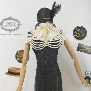 May include: A black beaded flapper dress with fringe detailing and a pearl necklace. The dress is on a mannequin with a black hat and feather.