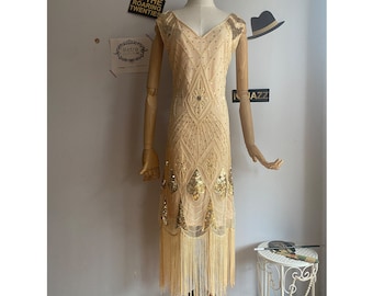 May include: A gold and beige flapper dress with fringe and sequins. The dress has a V-neckline and sleeveless design.