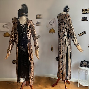 May include: A long black and gold sequined dress with a fringe hem. The dress has a high neckline and long sleeves. The dress is on a mannequin and is displayed against a white wall.