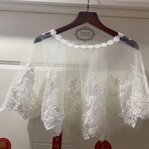 Peut inclure: Un châle en dentelle blanche transparente avec broderies florales, présenté sur un cintre en bois. Le châle présente des détails en dentelle délicate et un motif floral. Le mot "Retro" est visible sur le cintre.
