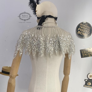 May include: A silver sequined cape with a scalloped edge. The cape is made of a sheer fabric and is decorated with small, silver sequins. The cape is a popular accessory for vintage-inspired fashion.