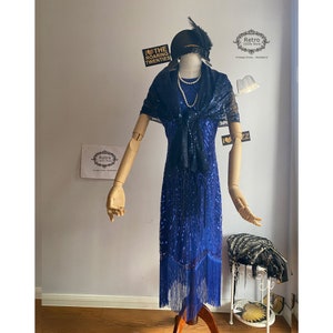 May include: A blue sequined flapper dress with fringe trim. The dress has a black lace shawl and a pearl necklace. The dress is on a mannequin with a black hat with a feather.