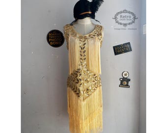 Peut inclure: Une robe de flapper pailletée dorée avec des détails en franges. La robe est sans manches et a un décolleté en V. La robe est sur un mannequin et a un bandeau noir avec une plume sur le dessus. Le texte "Retro 1920s Style" et "Vintage Dress - Headband" est visible sur l'image.
