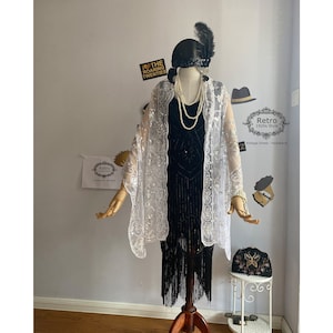 Könnte beinhalten: Ein weißer Spitzen-Schal mit silbernen Pailletten, getragen über einem schwarzen, perlenbesetzten Flapper-Kleid mit Fransen. Das Model trägt ein schwarzes Stirnband mit einer Feder und eine Perlenkette. Der Text "I ♥ THE ROARING TWENTIES" ist im Hintergrund sichtbar.