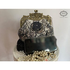 Pu&ograve; includere: Una pochette in satin argento con cornice in metallo dorato e ricamo floreale con perline nere. La pochette ha una tracolla a catena ed &egrave; appoggiata su una superficie bianca con un telefono nero e una collana di perle.
