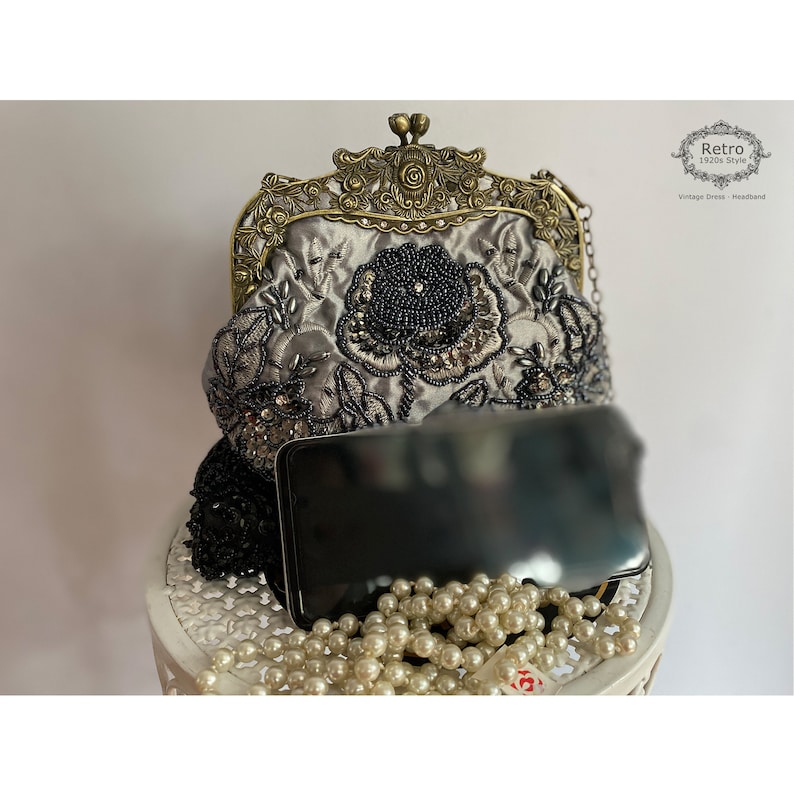 Pu&ograve; includere: Una pochette in satin argento con cornice in metallo dorato e ricamo floreale con perline nere. La pochette ha una tracolla a catena ed &egrave; appoggiata su una superficie bianca con un telefono nero e una collana di perle.
