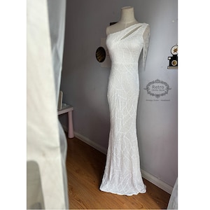 May include: A white one-shoulder beaded gown with a cut-out detail and fringe on the shoulder. The gown has a fitted bodice and a flowing skirt. The dress is on a mannequin in a room with a white wall and a wooden floor. The text "Retro 1920s Style Vintage Dress Headband" is visible in the lower right corner of the image.