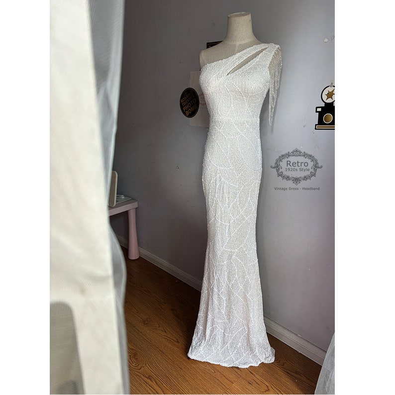 May include: A white one-shoulder beaded gown with a cut-out detail and fringe on the shoulder. The gown has a fitted bodice and a flowing skirt. The dress is on a mannequin in a room with a white wall and a wooden floor. The text "Retro 1920s Style Vintage Dress Headband" is visible in the lower right corner of the image.