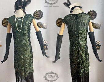 May include: A green sequined flapper dress with fringe trim and a matching headband. The dress has a short sleeve and a fitted bodice. The headband is adorned with feathers. The dress is styled on a mannequin with black gloves and a pearl necklace.