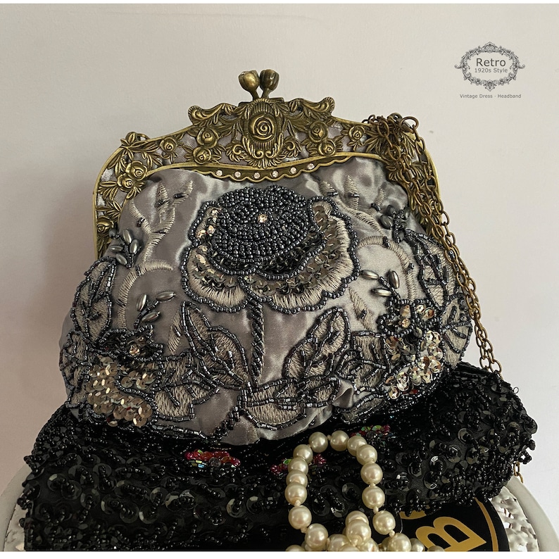 Pu&ograve; includere: Una borsa da sera in satin grigio con un design floreale e decorazioni di perline. La borsa ha una chiusura in ottone e una tracolla a catena.