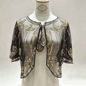 May include: A black sheer bolero jacket featuring intricate gold sequin embroidery in leaf and paisley patterns. The garment has short sleeves, a rounded neckline, and a front closure with a single decorative sequin-embellished tab.