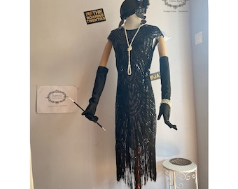 May include: A black sequined flapper dress with fringe detailing. The dress is on a mannequin wearing a black hat with a feather and black gloves. The mannequin is standing on a wooden stand. The dress has a beaded necklace and a pearl bracelet.