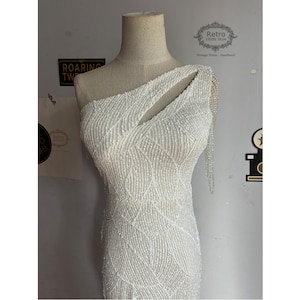 May include: A white one-shoulder sequined dress with a cut-out detail on the shoulder. The dress has a fitted silhouette and a geometric pattern.