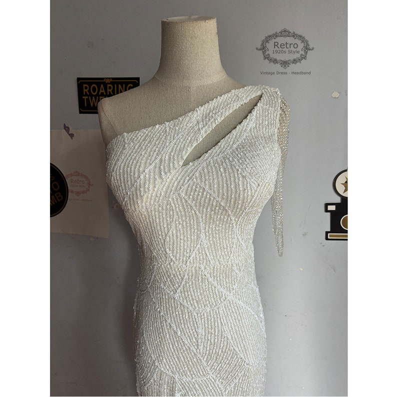 May include: A white one-shoulder sequined dress with a cut-out detail on the shoulder. The dress has a fitted silhouette and a geometric pattern.